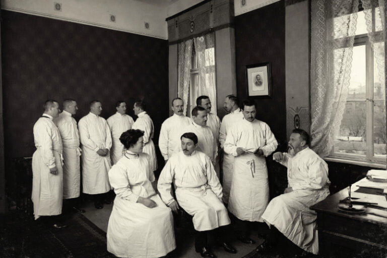 Politische Verfolgung in Deutschland 31 Schwarzweißes historisches Foto von Rahel Hirsch in einer Gruppe von Ärzten in weißen Kitteln, die in einem Arbeitszimmer um 1900 miteinander sprechen und sitzen; im Raum stehen ein Schreibtisch mit Papieren, hohe Fenster mit Spitzengardinen und dunkle Wandvertäfelungen.
