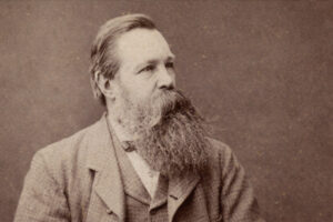 Porträt von Friedrich Engels im Sepia-Ton im 16:9-Format, mit langem Bart und Anzug, nach rechts blickend vor neutralem Hintergrund.