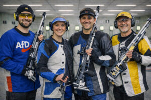 Vier Sportschützen mit Luftgewehren in einer Indoor-Schießanlage, links ein Schütze mit Jacke und AfD-Logo
