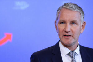 Porträt von Björn Höcke im Anzug mit grauem Haar und blauem Hintergrund mit AfD-Logo der leicht nach links blickt.