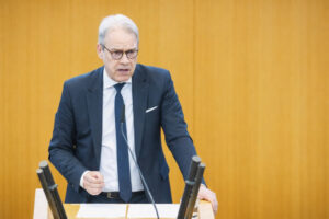 Georg Maier mit Brille und dunkelblauem Anzug spricht an einem Rednerpult mit Mikrofonen vor einer gelben Wand.