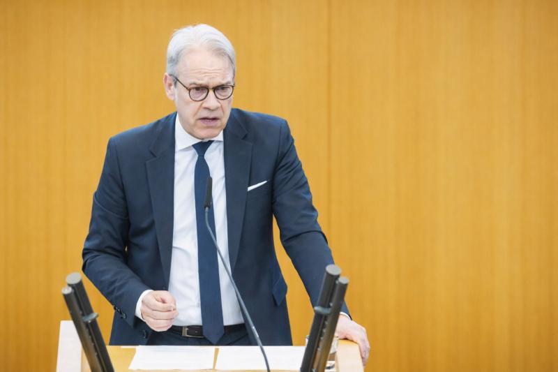 Georg Maier mit Brille und dunkelblauem Anzug spricht an einem Rednerpult mit Mikrofonen vor einer gelben Wand.