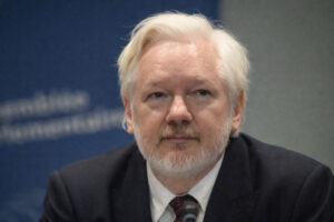 Nahaufnahme von weißhaarigen Julian Assange mit Bart in dunklem Anzug und Krawatte, der bei einer formellen Anhörung vor einem Mikrofon sitzt; unscharfer blauer Hintergrund mit Schrift.