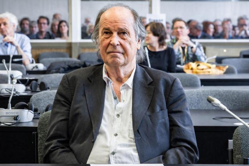 Dr Walter Weber, dressed in a suit, is sitting in the courtroom in front of the microphones; in the background, the defendants are seated in the dock.