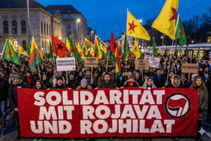 Großer Protestzug bei Dämmerung in einer deutschen Innenstadt. Teilnehmer halten ein großes rotes Banner mit der weißen Aufschrift 'SOLIDARITÄT MIT ROJAVA UND ROJHILAT'. Im Hintergrund wehen kurdische YPG-Flaggen mit rotem Stern und gelbem Grund sowie Antifa-Flaggen. Ein Straßenbahngleis und historische Gebäude sind sichtbar.
