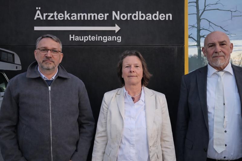 Monika Jiang mit ihren beiden Anwälten vor einem Gebäude mit der Aufschrift „Ärztekammer Nordbaden“ und dem Hinweis „Haupteingang“ mit Pfeil nach rechts.