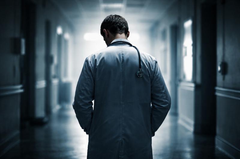 Deprimierter Arzt von hinten in einem dunklen Krankenhausflur, mit gesenktem Kopf und Stethoskop, symbolisiert Belastung und Isolation im medizinischen Alltag