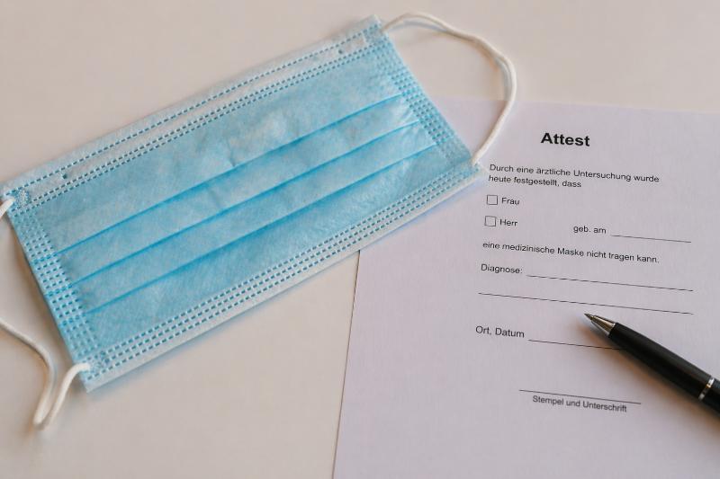 Maskenatteste: Blaue Einweg-Gesichtsmaske neben einem ärztlichen Attestformular und Stift auf hellem Hintergrund