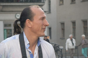 Peter Fitzek mit Pferdeschwanz und Kamera um den Hals blickt seitlich in einer städtischen Umgebung mit unscharfen Passanten im Hintergrund.