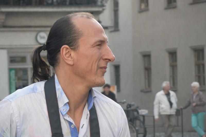 Peter Fitzek mit Pferdeschwanz und Kamera um den Hals blickt seitlich in einer städtischen Umgebung mit unscharfen Passanten im Hintergrund.