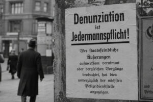 Historisch anmutendes Schwarzweißbild mit einem Plakat „Denunziation ist Jedermannspflicht!“ an einer Häuserwand in einer deutschen Straßenszene.