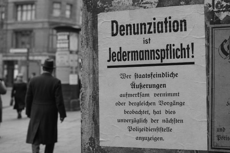 Historisch anmutendes Schwarzweißbild mit einem Plakat „Denunziation ist Jedermannspflicht!“ an einer Häuserwand in einer deutschen Straßenszene.