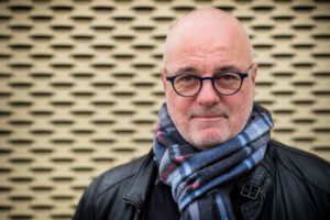 Porträtbild von Stefan Hockertz mit Brille, Schal und Lederjacke vor strukturiertem Hintergrund im Querformat