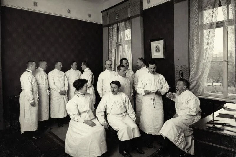 Schwarzweißes historisches Foto von Rahel Hirsch in einer Gruppe von Ärzten in weißen Kitteln, die in einem Arbeitszimmer um 1900 miteinander sprechen und sitzen; im Raum stehen ein Schreibtisch mit Papieren, hohe Fenster mit Spitzengardinen und dunkle Wandvertäfelungen.