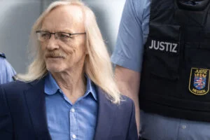 Maximilian Eder mit langen grauen Haaren, Brille, blauem Hemd und Sakko wird von Justizbeamten begleitet.