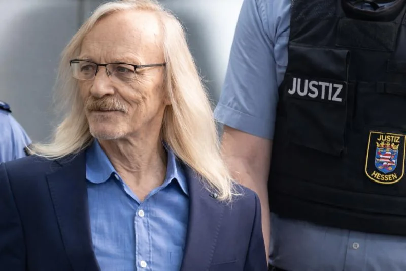 Maximilian Eder mit langen grauen Haaren, Brille, blauem Hemd und Sakko wird von Justizbeamten begleitet.
