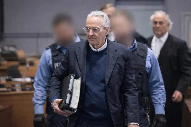 Rüdiger von Pescatore mit grauen Haaren und Brille trägt Aktenordner und wird von Justizbeamten in einem Gerichtsgebäude begleitet.