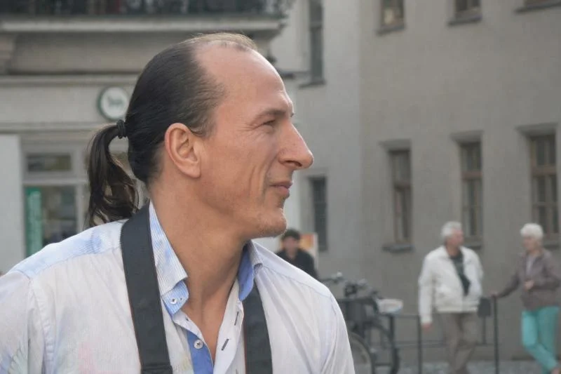 Peter Fitzek und das Königreich Deutschland (KRD): Zwischen Staatsutopie und Justizrepression 1 Peter Fitzek mit Pferdeschwanz und Kamera um den Hals blickt seitlich in einer städtischen Umgebung mit unscharfen Passanten im Hintergrund.